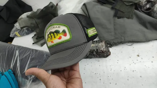 Gorra trucker gris con malla negra, parche de pavón y etiqueta lateral