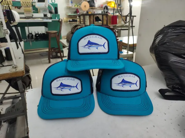 Tres gorras trucker turquesa con parche de marlín azul, con la bordadora y las máquinas de coser del taller al fondo